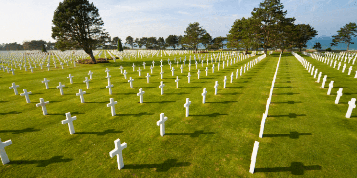 Rows of white crosses mark graves in a large, well-maintained cemetery with green grass, trees, and a view of the sea in the background.