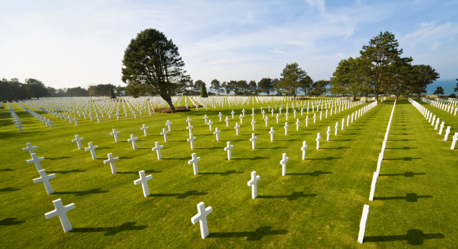 Rows of white crosses mark graves in a military cemetery, with green grass and scattered trees under a partly cloudy sky.