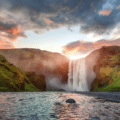 A wide waterfall cascades between green cliffs under a dramatic sunset sky, with a river in the foreground and a person standing near the falls.