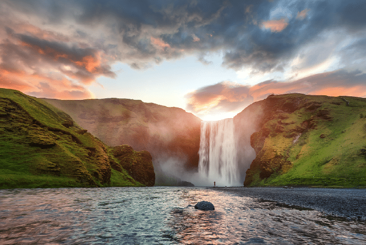 A wide waterfall cascades between green cliffs under a dramatic sunset sky, with a river in the foreground and a person standing near the falls.