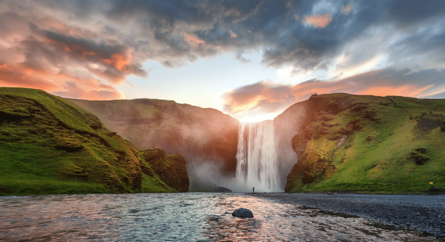 Wide waterfall cascading over a cliff between green hills at sunset, with a person standing near the base and water flowing in the foreground.
