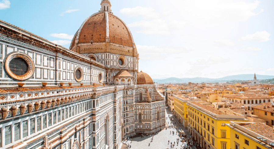 View of Florence Cathedral with its large dome, detailed marble façade, and surrounding city buildings under a bright, clear sky. People are visible walking in the square below.