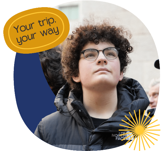 A young person with curly hair and glasses looks upwards while wearing a dark puffer jacket. Text reads "Your trip, your way" with a yellow sunburst graphic in the corner.