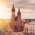 A large brick church with two uneven towers stands in a city square surrounded by historic buildings under a cloudy sky with sunlight.