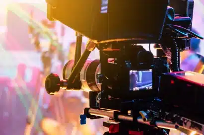 Close-up of a professional video camera on a tripod, with colourful lights and a blurred background.