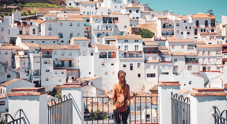 A person with a rucksack stands on a terrace, facing a hillside covered with tightly packed white buildings under a clear blue sky.