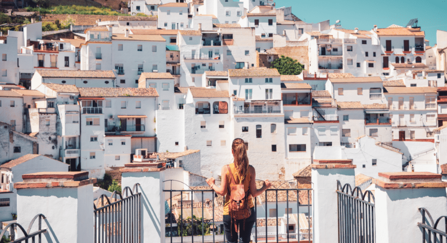 A person with a rucksack stands on a balcony, looking at a hillside covered with white buildings under a clear blue sky.