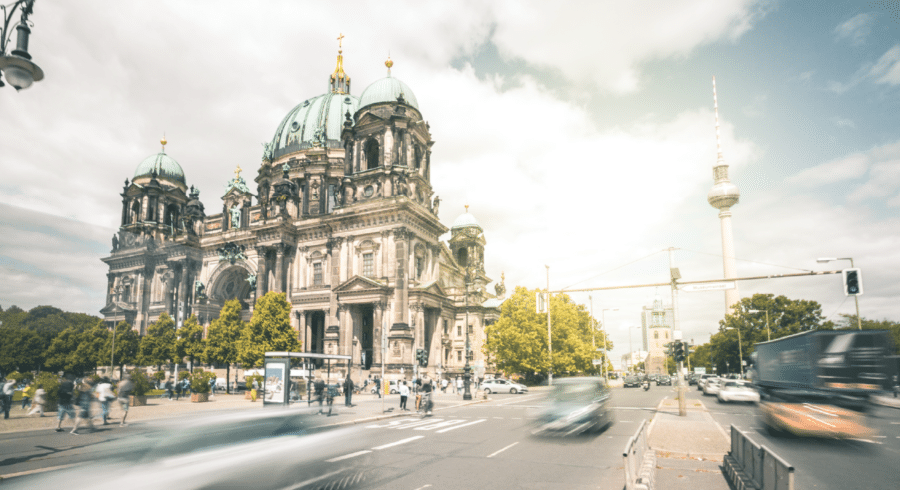 View of Berlin Cathedral with the Fernsehturm TV tower in the background, cars and pedestrians moving along the street in the foreground on a partly cloudy day.