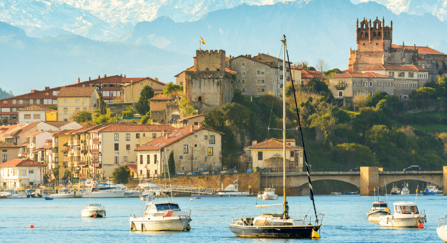 Boats on water in front of a historic town with stone buildings, a castle, and a church, set against a backdrop of mountains.