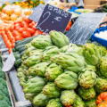 Artichokes, broccoli, tomatoes, and other vegetables displayed for sale at an outdoor market with handwritten price signs.