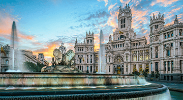 The photo shows the Cibeles Fountain in front of the Palacio de Cibeles in Madrid at sunset, with water jets and a partly cloudy sky.