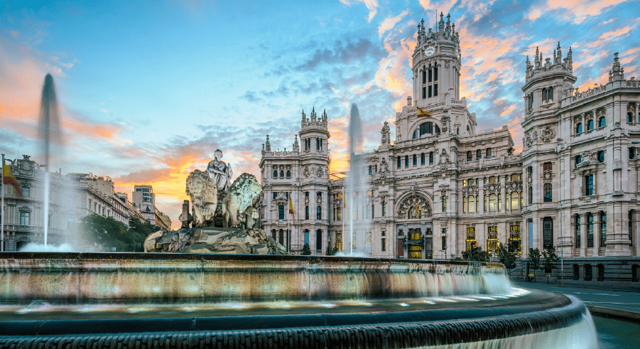 The image shows the Cibeles Fountain and the Palacio de Cibeles in Madrid, Spain, with a colourful sky at sunset in the background.