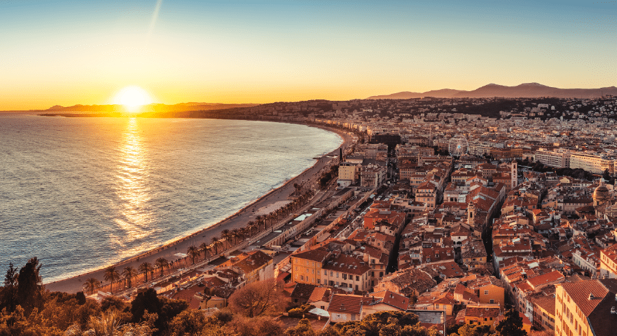 Aerial view of a coastal city at sunset with the sun near the horizon, a long beach, and buildings stretching along the shoreline.