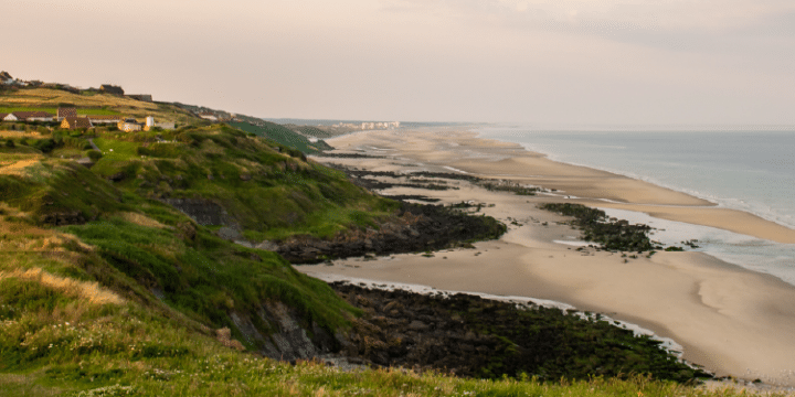 A coastal landscape with grassy cliffs, scattered houses, and a sandy beach with rocks stretching along the shoreline under a cloudy sky.