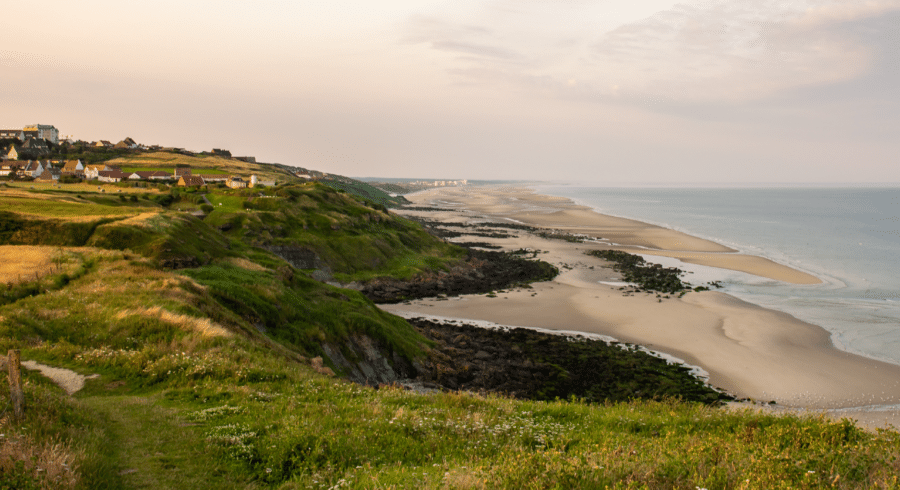 Coastal landscape with grassy cliffs, scattered houses, and a wide sandy beach stretching along the shoreline under a partly cloudy sky.