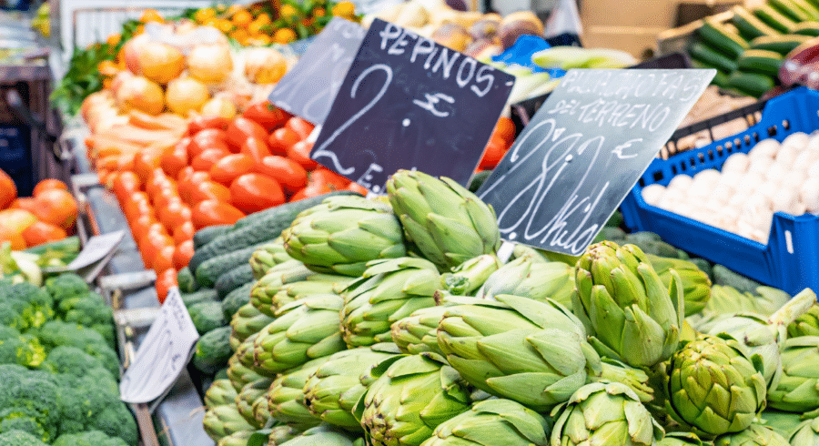 Assorted fresh vegetables, including artichokes, tomatoes, cucumbers, and broccoli, displayed at a market stall with handwritten price signs.