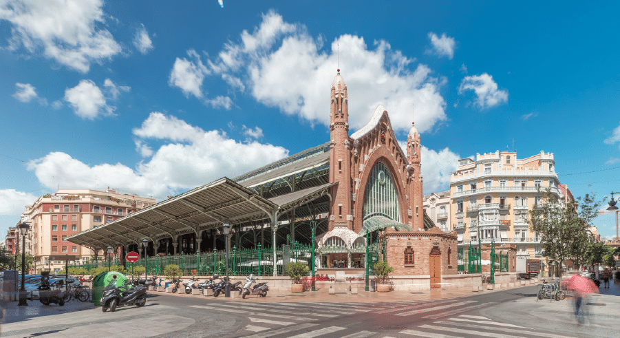 A large historic market building with a glass roof and brick entrance stands in a city square under a blue sky with scattered clouds.