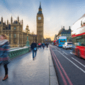 People walk across Westminster Bridge in London, with Big Ben and the Houses of Parliament in the background and a red double-decker bus passing by.