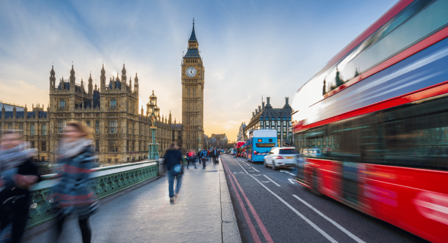 Pedestrians and vehicles, including a red double-decker bus, move along Westminster Bridge in London with Big Ben and the Houses of Parliament in the background at sunset.