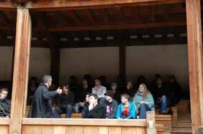 A group of people sit on wooden benches under a roof whilst a person in front speaks to them, likely giving a talk or lesson.