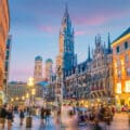 Evening view of Marienplatz in Munich, Germany, with the New Town Hall and Frauenkirche towers, surrounded by colourful buildings and people walking.