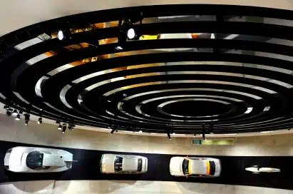 Overhead view of an exhibition with five cars displayed under a large, circular black spiral structure with spotlights.