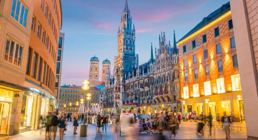 City square at dusk in Munich, Germany, with people walking and the illuminated New Town Hall and Frauenkirche towers in the background.
