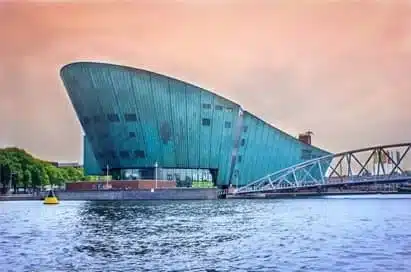 A modern, green, boat-shaped building stands by the waterfront—an ideal spot for science trips. Nearby, a bridge and trees complement the scene under a pastel sky reminiscent of Iceland’s iconic landscapes.