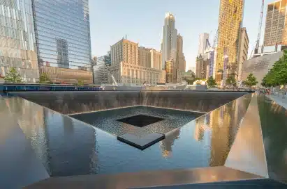 Reflecting pool with waterfalls at the 9/11 Memorial in New York City, surrounded by tall buildings under clear sky.