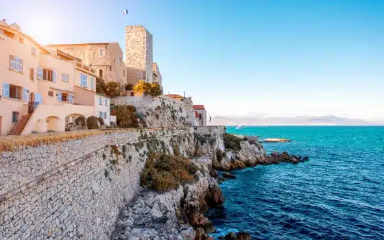Stone buildings and a fortress sit atop a rocky coastline overlooking the blue sea under a clear sky.