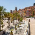 Palm-lined promenade along a sandy beach with colourful hillside buildings and people enjoying the sunny day.