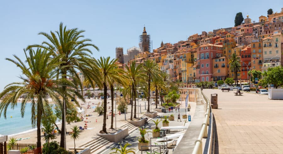 A seaside promenade lined with palm trees, people on the sandy beach, and colourful hillside buildings under a clear blue sky.