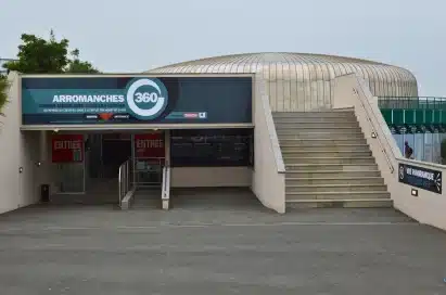 Entrance to Arromanches 360, a circular theatre with a modern dome, showing signs for tickets and entry, with stairs leading to an upper level.
