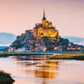 Mont Saint-Michel, an island commune with a historic abbey, is illuminated at sunset, surrounded by water and tidal flats in Normandy, France.