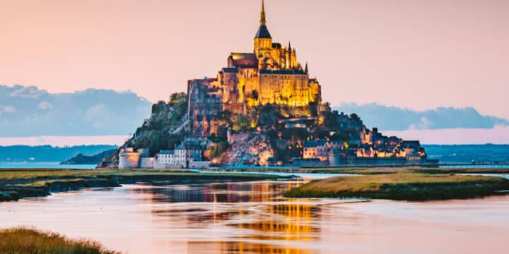 Mont Saint-Michel, an island commune with a historic abbey, is illuminated at sunset, surrounded by water and tidal flats in Normandy, France.