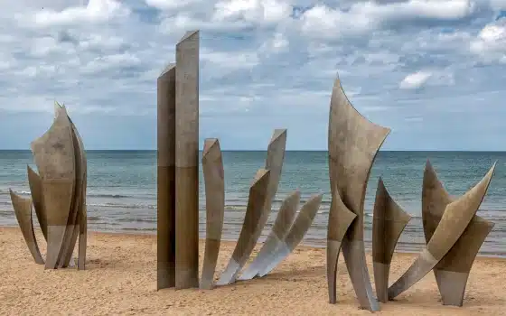 Metallic sculpture with tall, curved panels stands on a sandy beach with the sea and cloudy sky in the background.