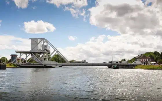 A metal drawbridge spans a wide river under a partly cloudy sky, with buildings and trees visible in the background.