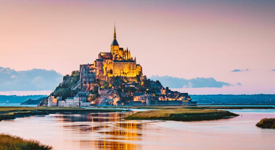 Mont Saint-Michel, a historic abbey and island commune in France, is illuminated at sunset, with its reflection visible in the calm water below.