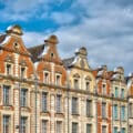 Row of ornate historic European buildings with red brick and stone facades, arched windows, and decorative gables under a partly cloudy sky.