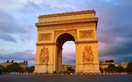 The Arc de Triomphe stands in an open square in Paris at sunset, with a partly cloudy sky in the background.