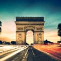 The Arc de Triomphe in Paris at sunset, with blurred headlights and taillights from passing cars on the road in the foreground.