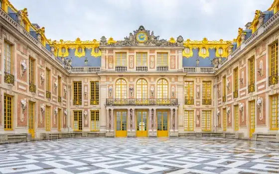 The courtyard of the Palace of Versailles features ornate architecture, golden accents, large windows, and a patterned black-and-white marble floor under a cloudy sky.