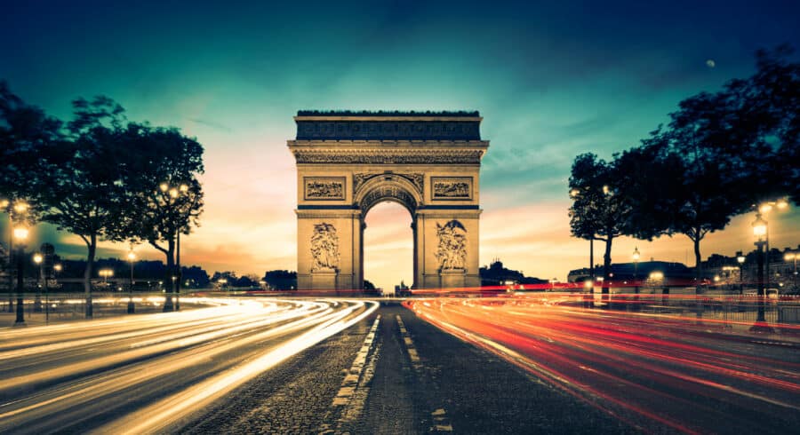 The Arc de Triomphe in Paris at dusk, with car light trails streaming past on either side under a colourful sky.