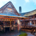 A wooden open-air theatre with a thatched roof, tiered seating, and a central stage, resembling the historic Globe Theatre in London.
