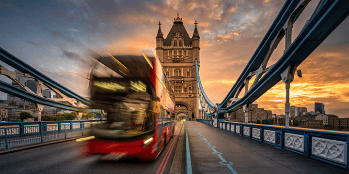 A red double-decker bus crosses Tower Bridge in London at sunset, with the sky displaying dramatic clouds and warm colours.