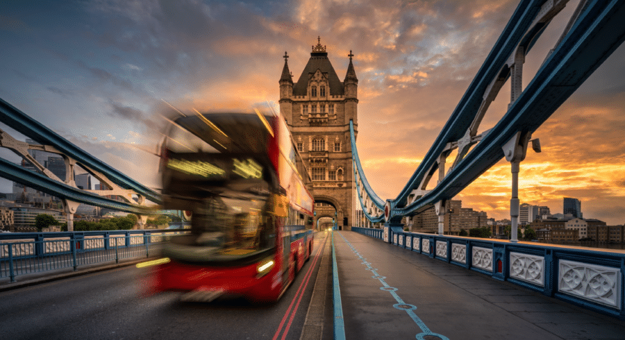 A red double-decker bus crosses Tower Bridge in London at sunset, with the bridge's towers and dramatic sky in the background.