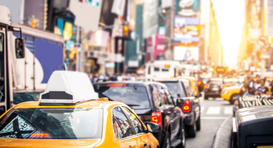 Traffic congestion with yellow taxis and cars on a busy city street during daylight, with tall buildings and hoardings visible in the background.