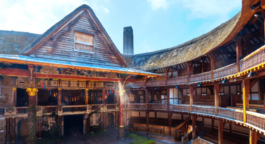 A view of the wooden stage and tiered seating inside Shakespeare’s Globe Theatre in London, with sunlight streaming in under a partly clear sky.
