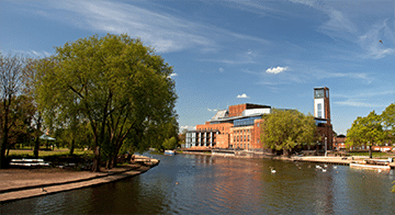 A river flows beside a red-brick building with a tall tower, surrounded by trees and a clear blue sky.