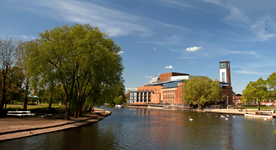 A red-brick theatre building sits by a river with ducks and swans, surrounded by green trees and a park, under a blue sky with scattered clouds.
